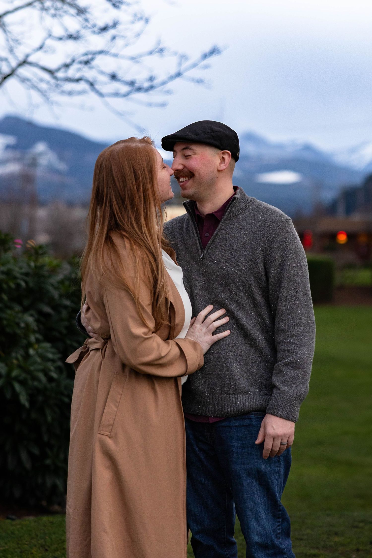 Couple sharing a kiss outdoors with mountain backdrop, woman in beige coat and man in grey sweater and flat cap.