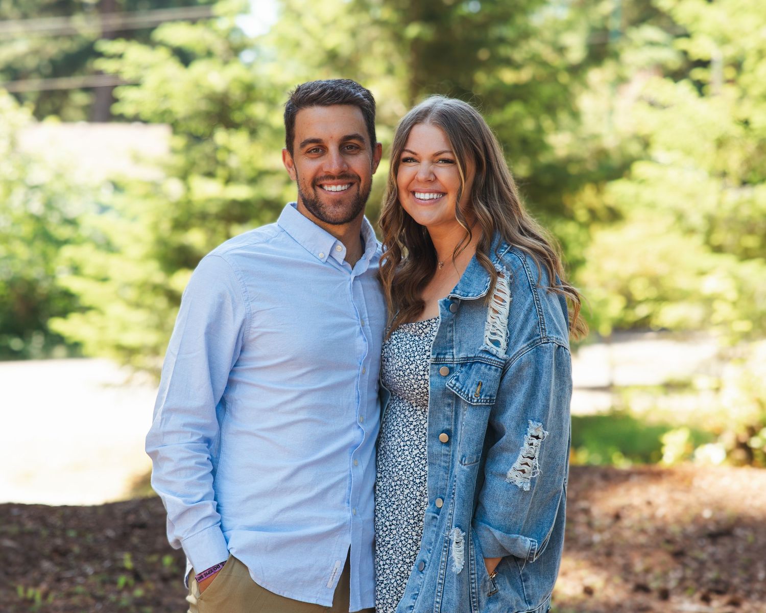 Smiling couple poses outdoors among green trees, man in blue shirt and woman in floral dress with denim jacket.