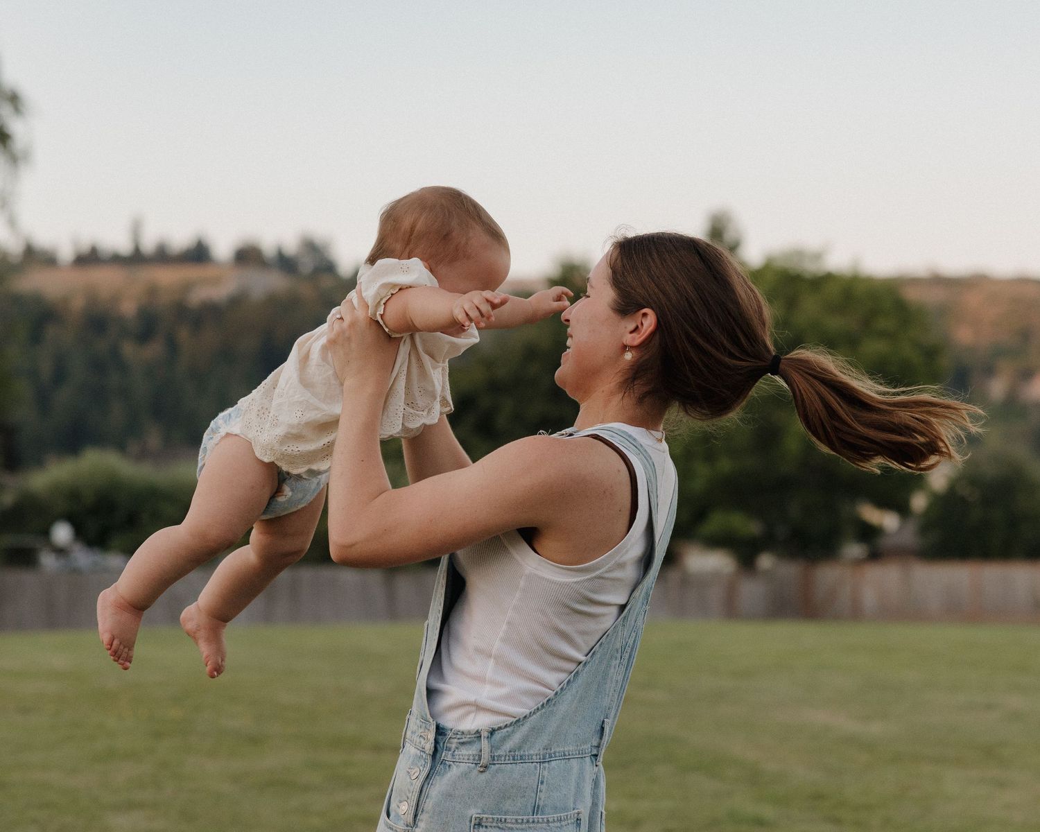 Young mother in denim overalls lifting happy baby outdoors in green field at sunset.