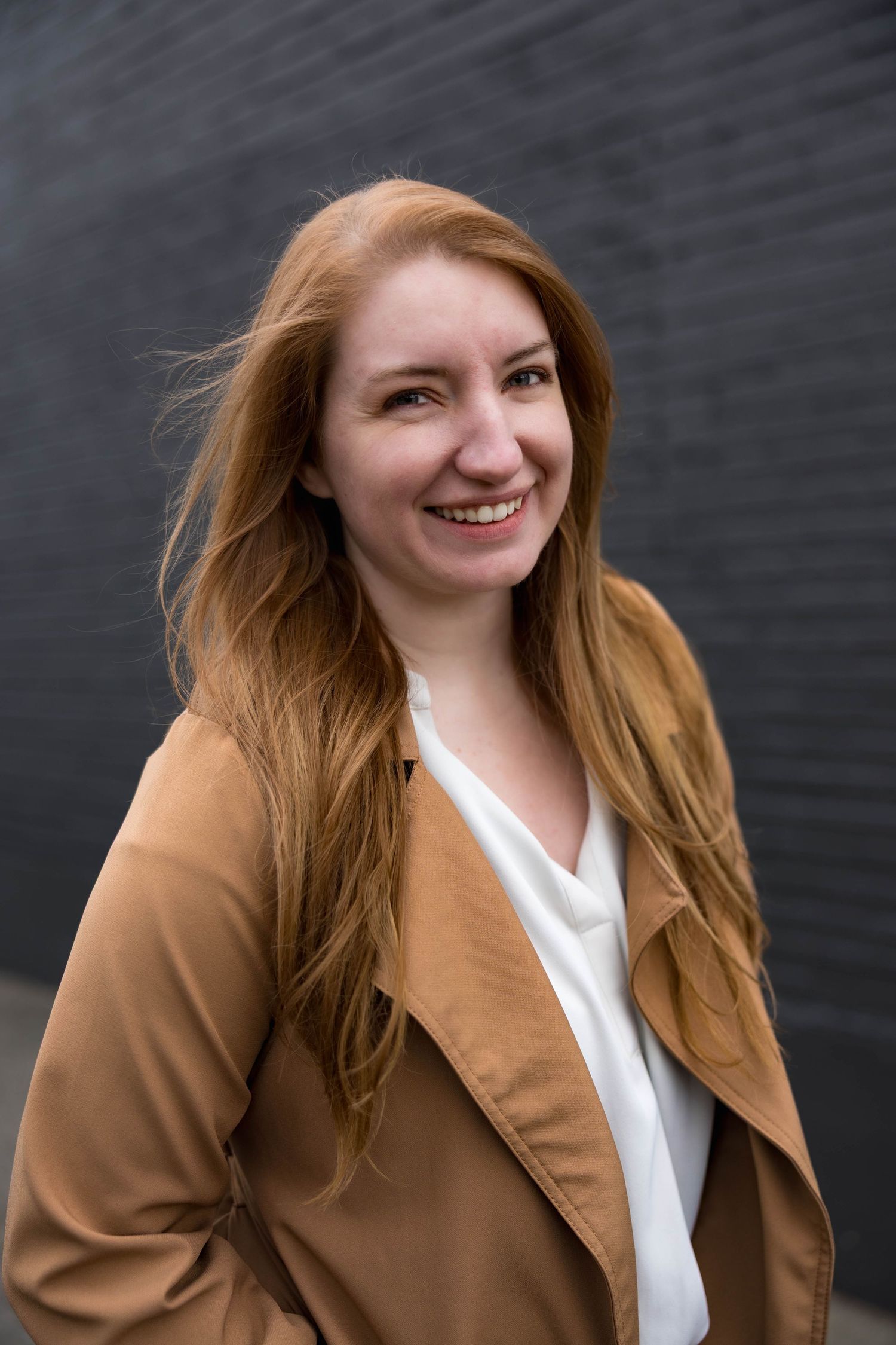 Cheerful young woman with long red hair in a tan jacket smiling outdoors.
