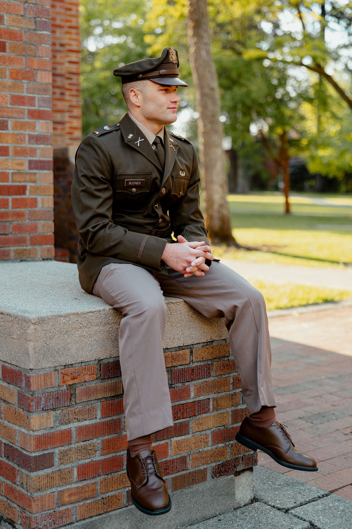 Military officer in dress uniform sitting on brick wall in campus courtyard.