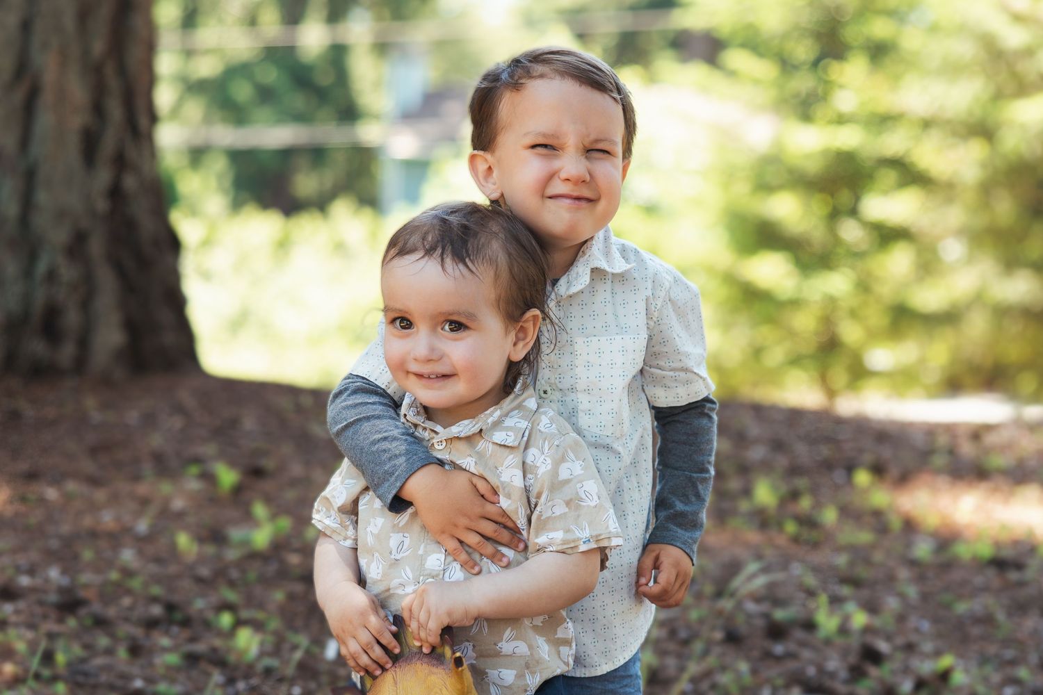 Two young brothers hugging and smiling together in a sunlit outdoor park setting.