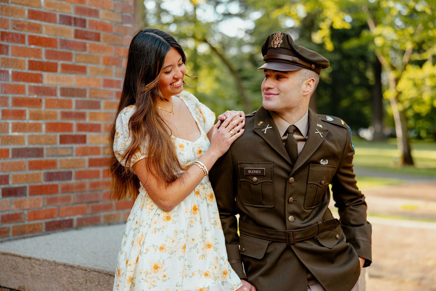 Happy couple sharing a candid moment by brick wall, woman in floral dress touching mans military uniform lapel.