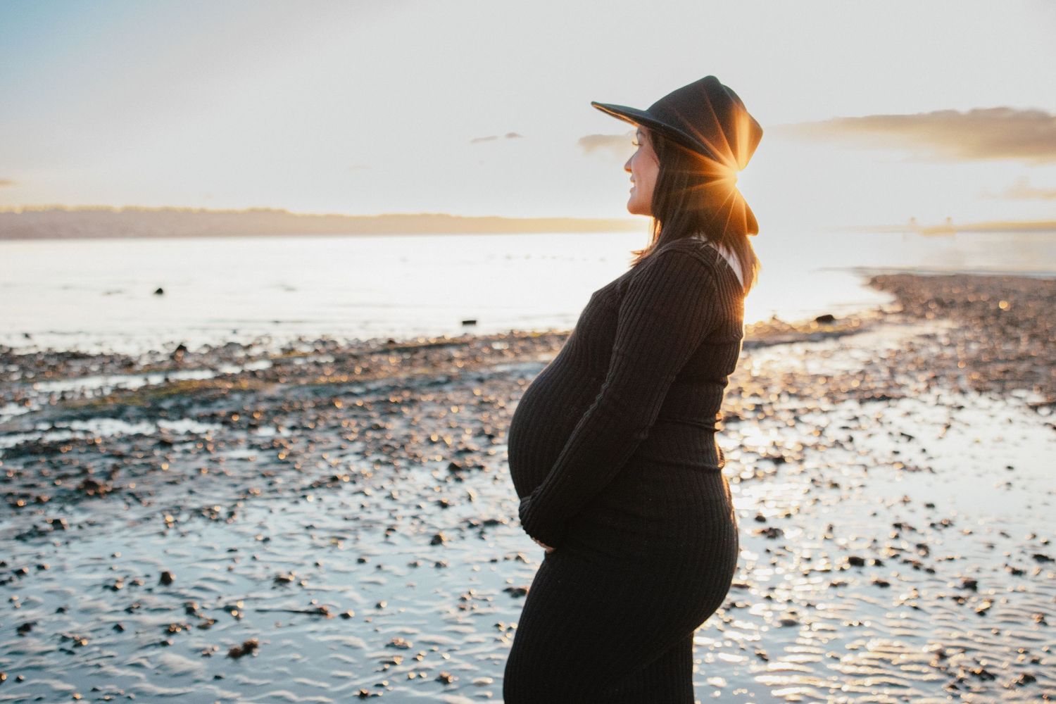 Pregnant woman in black dress and wide-brim hat cradling belly on a rocky beach at sunset.