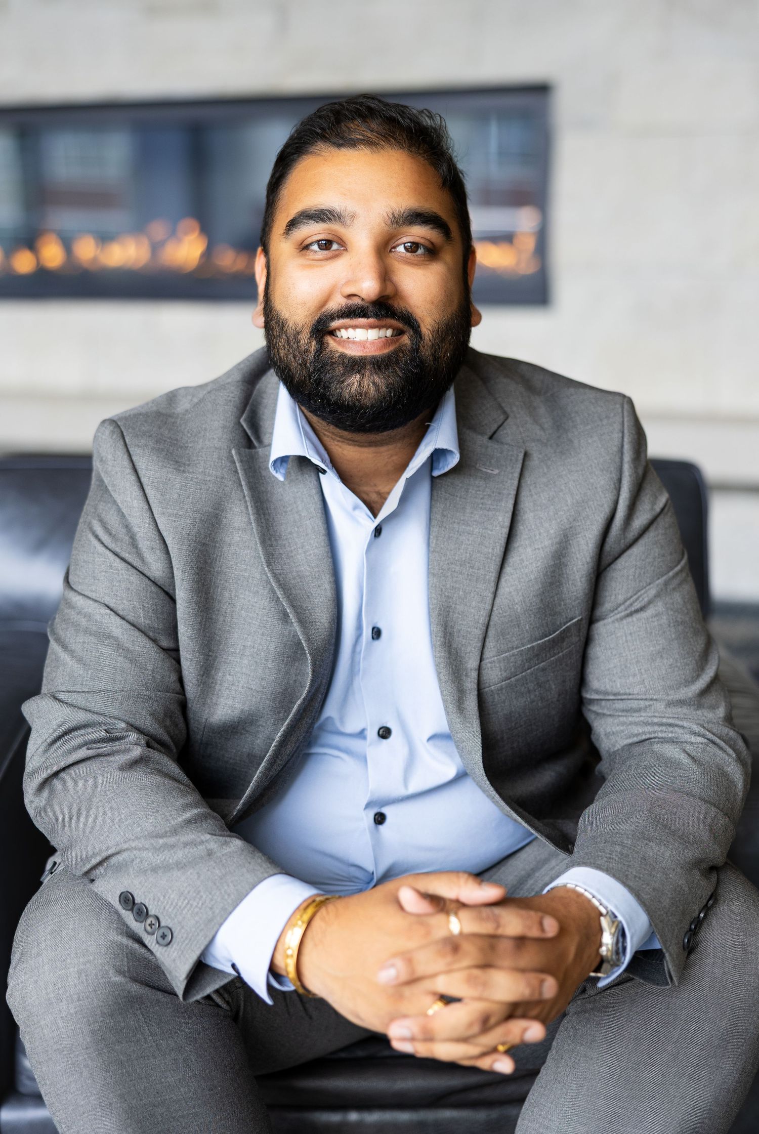 Bearded South Asian man in gray suit and blue shirt seated smiling in modern office with fireplace.