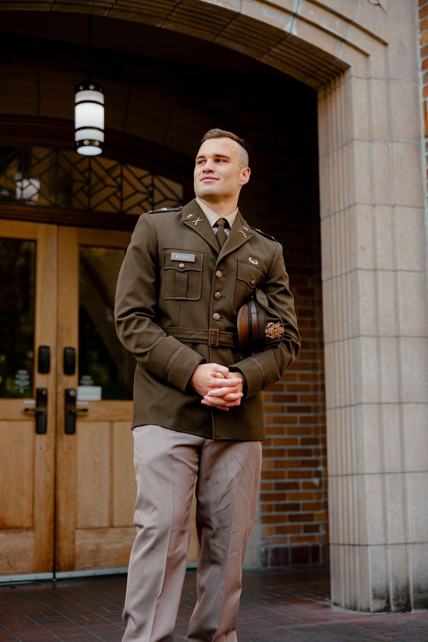 Young military officer in dress uniform standing confidently outside an arched stone building entrance.