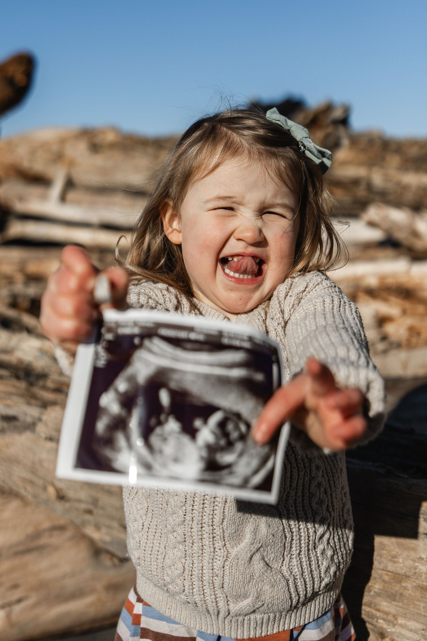 Excited toddler girl holding ultrasound photos outdoors, laughing with mouth open wide.