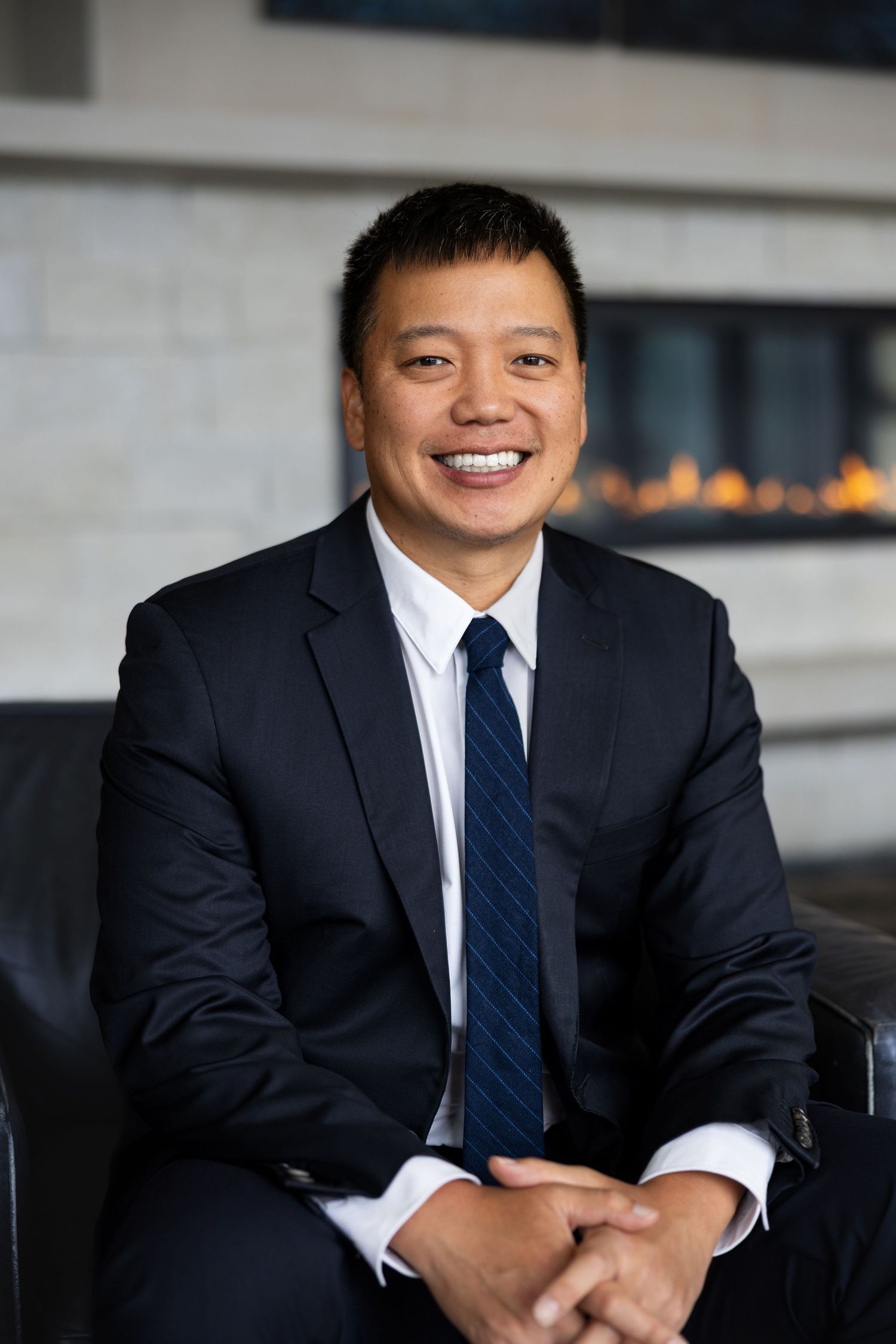 Smiling Asian man in dark navy suit and striped tie seated in modern office with fireplace background.