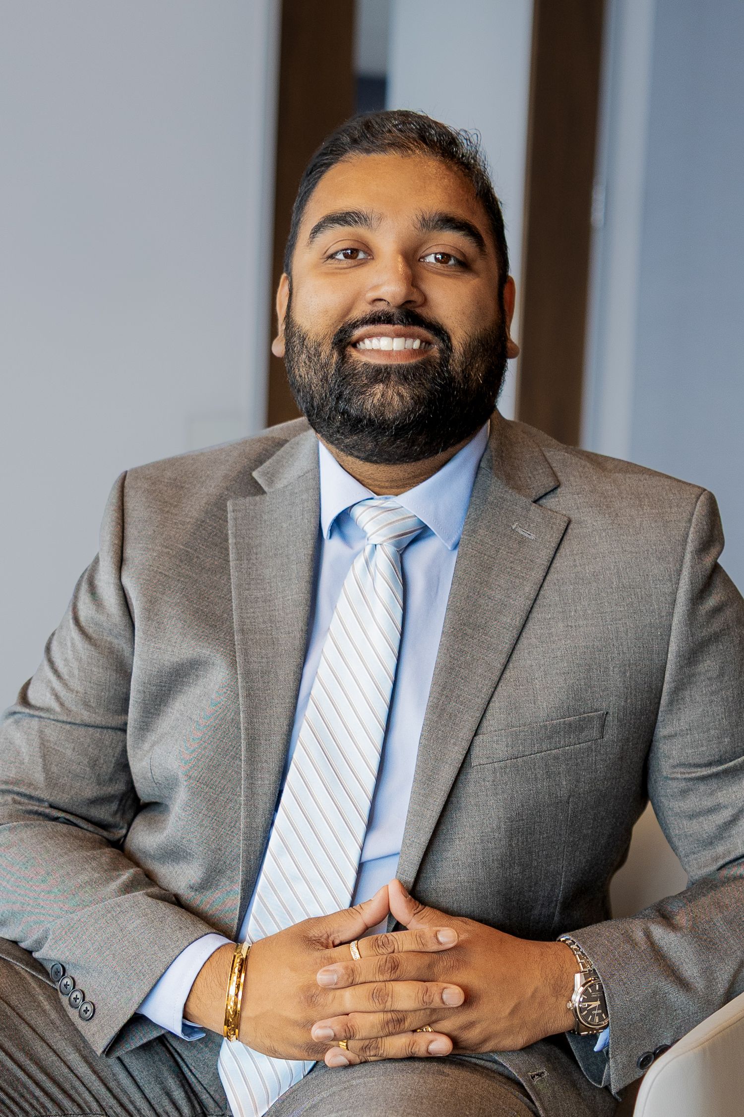 Bearded South Asian man in gray suit and striped tie seated, smiling warmly in modern office setting.