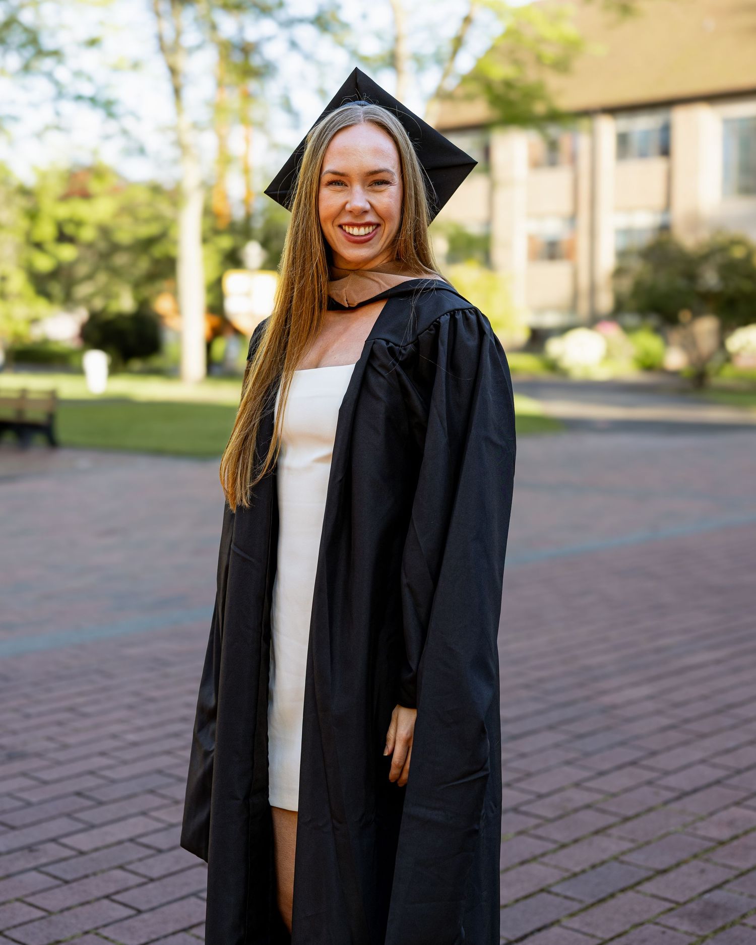 Graduate smiling in black cap and gown on sunny university campus plaza.