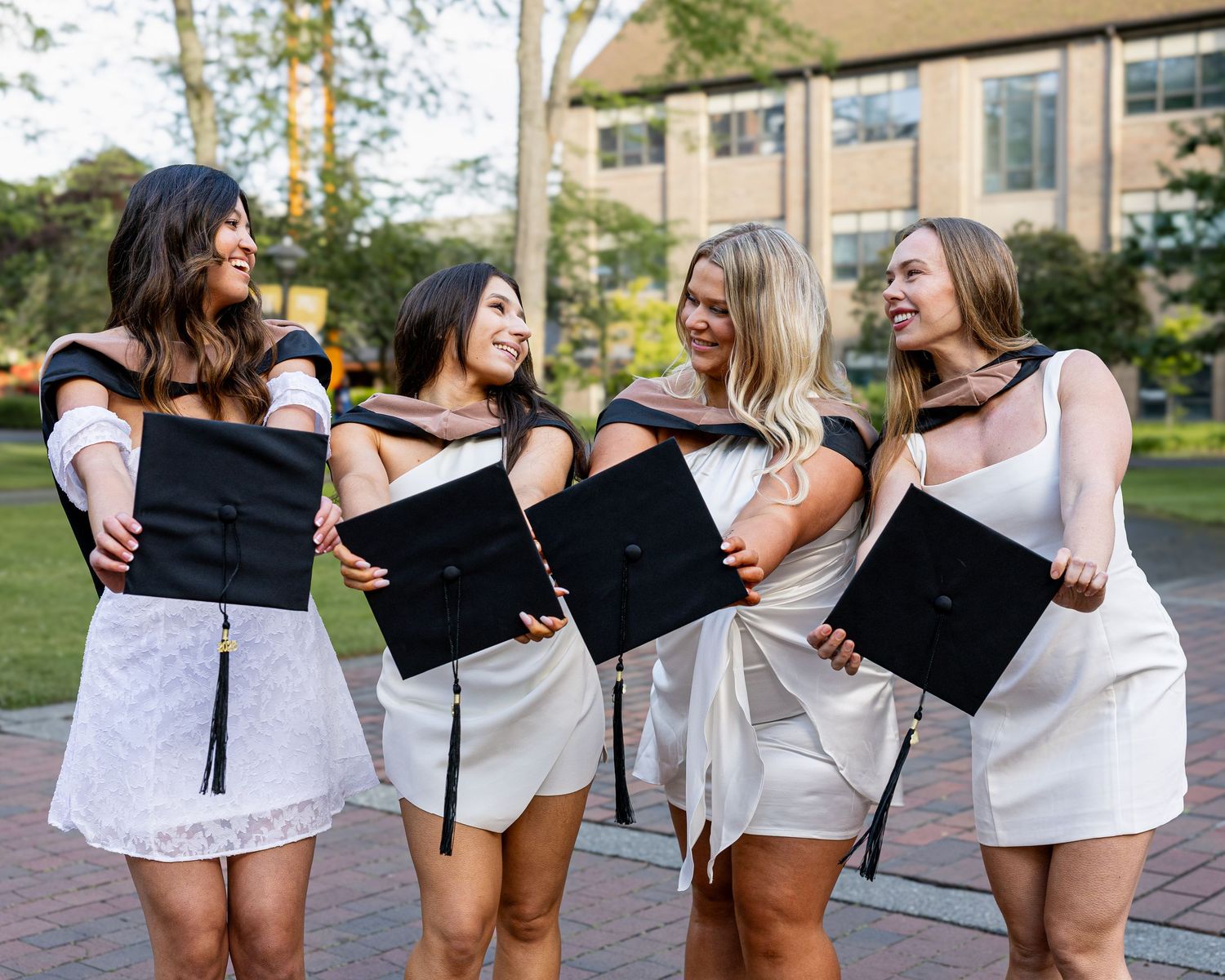 Four smiling female graduates in white dresses hold black graduation caps while posing together on a college campus.