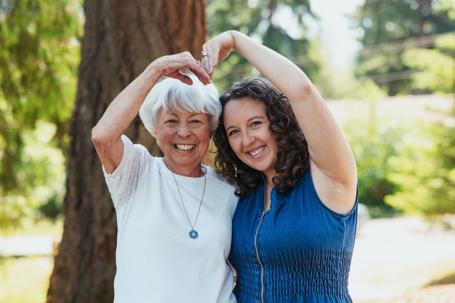 Two women making a heart shape with their arms raised outdoors in a park.