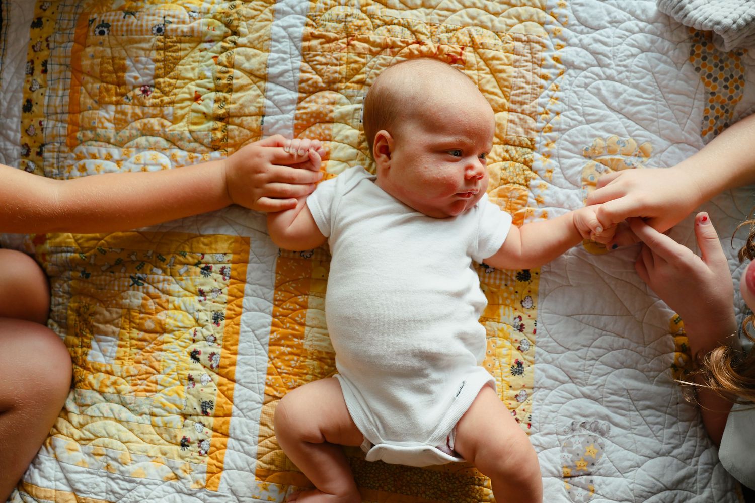 Newborn baby in white onesie lying on yellow quilt while two people gently hold its hands.