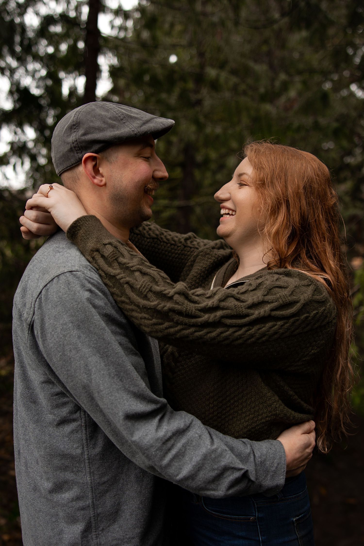 Laughing redhead woman hugs man in gray cap and shirt outdoors in a forest setting.