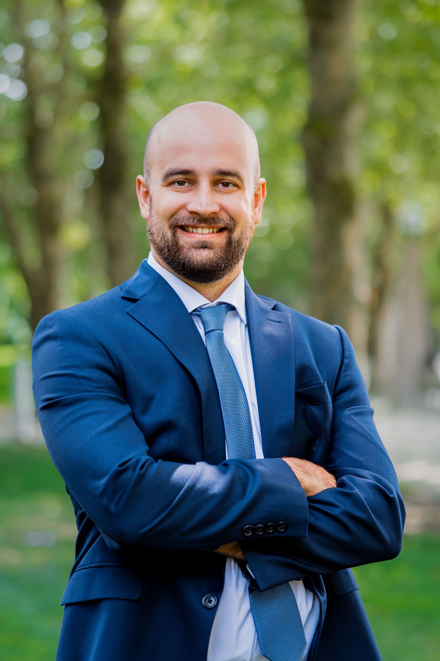 Confident bald bearded man in navy suit standing arms crossed smiling in a park setting.