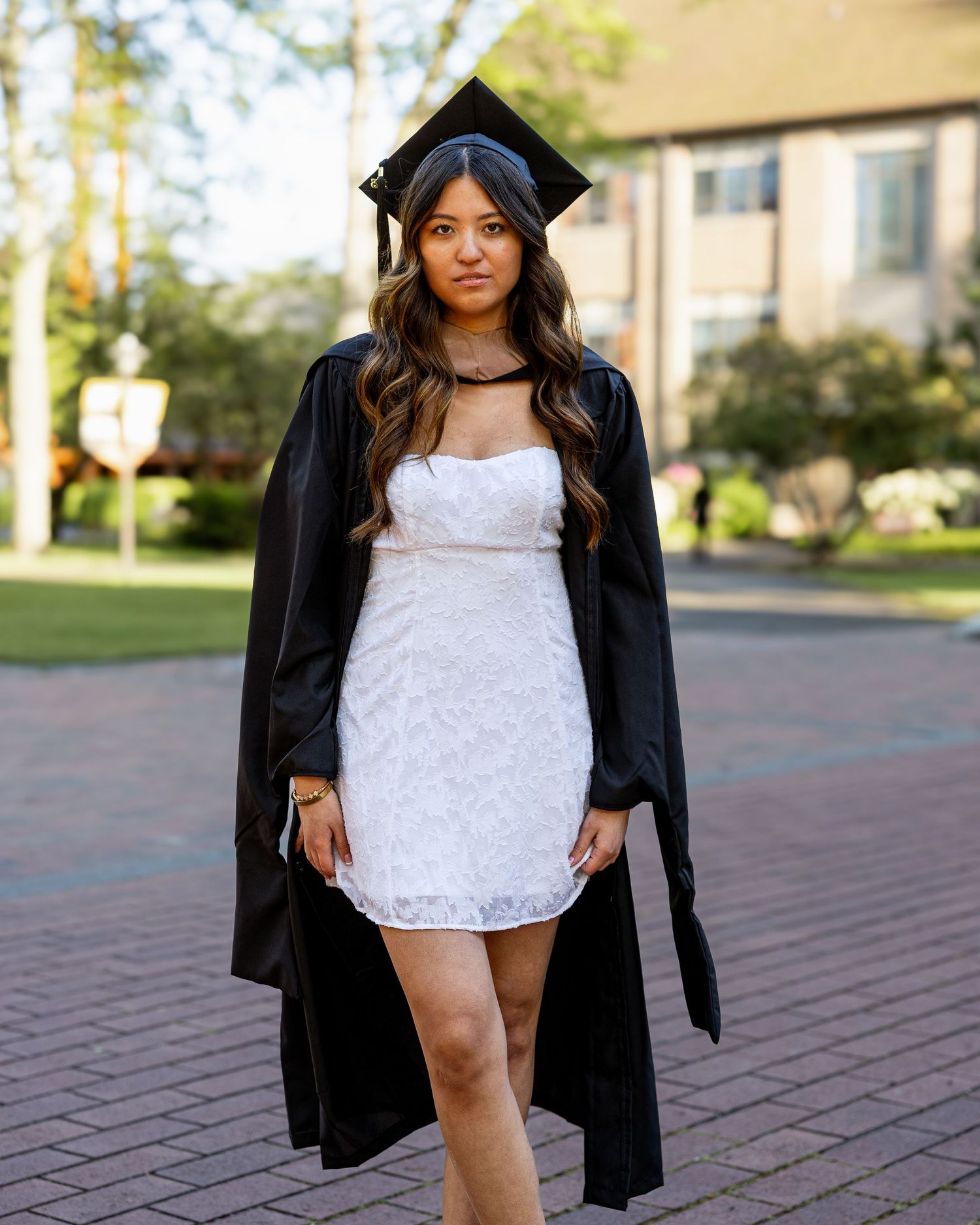 Female graduate in white lace mini dress and black cap and gown walks on a brick college campus path.
