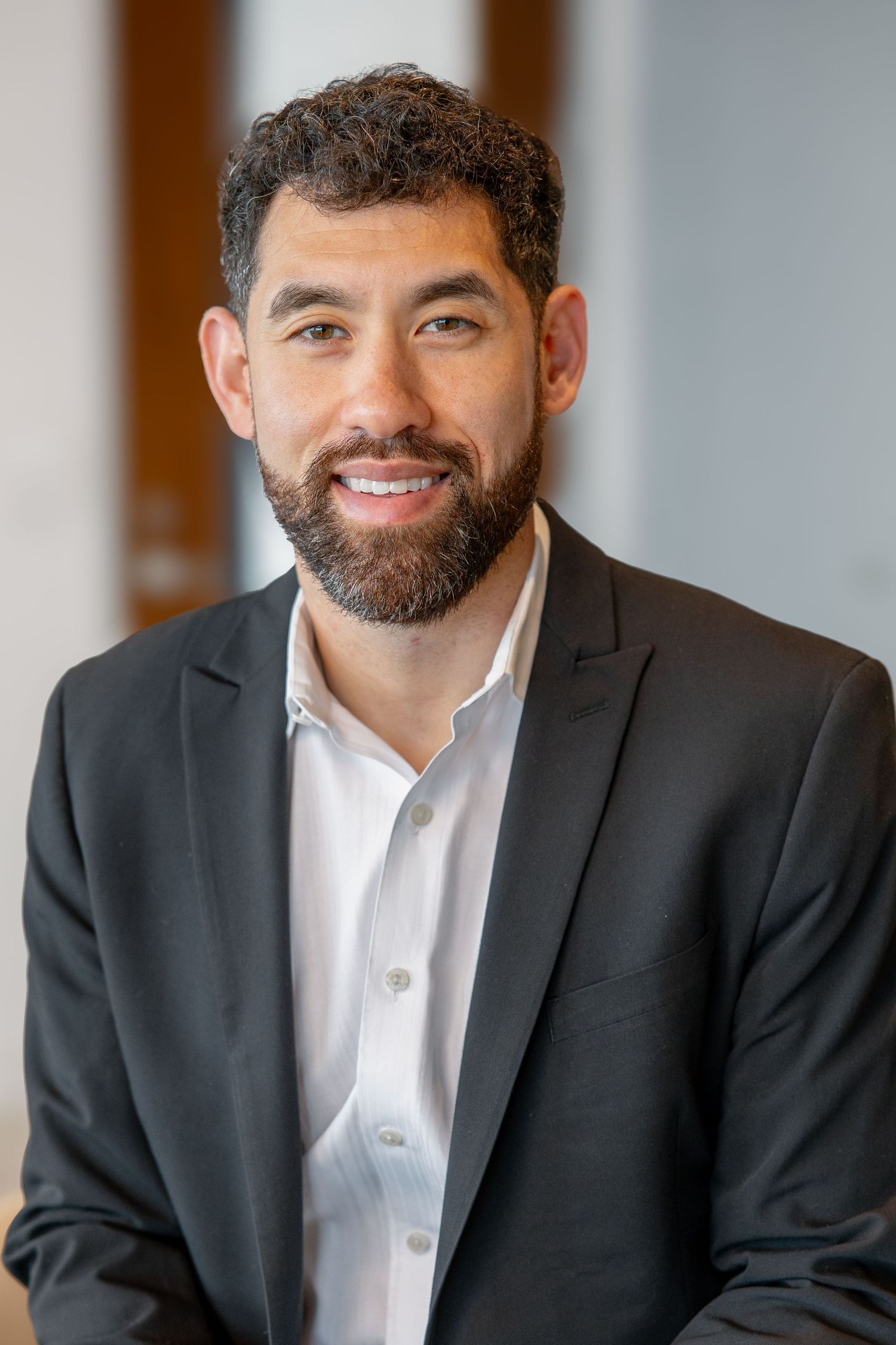Bearded businessman in dark blazer smiling confidently in a bright professional setting.