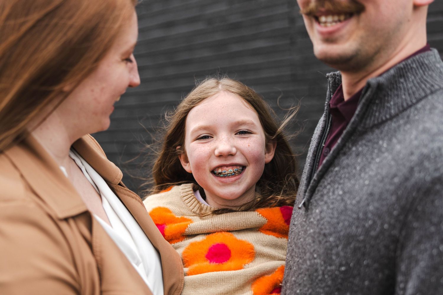 Smiling girl with braces wrapped in colorful floral blanket between laughing parents outdoors.