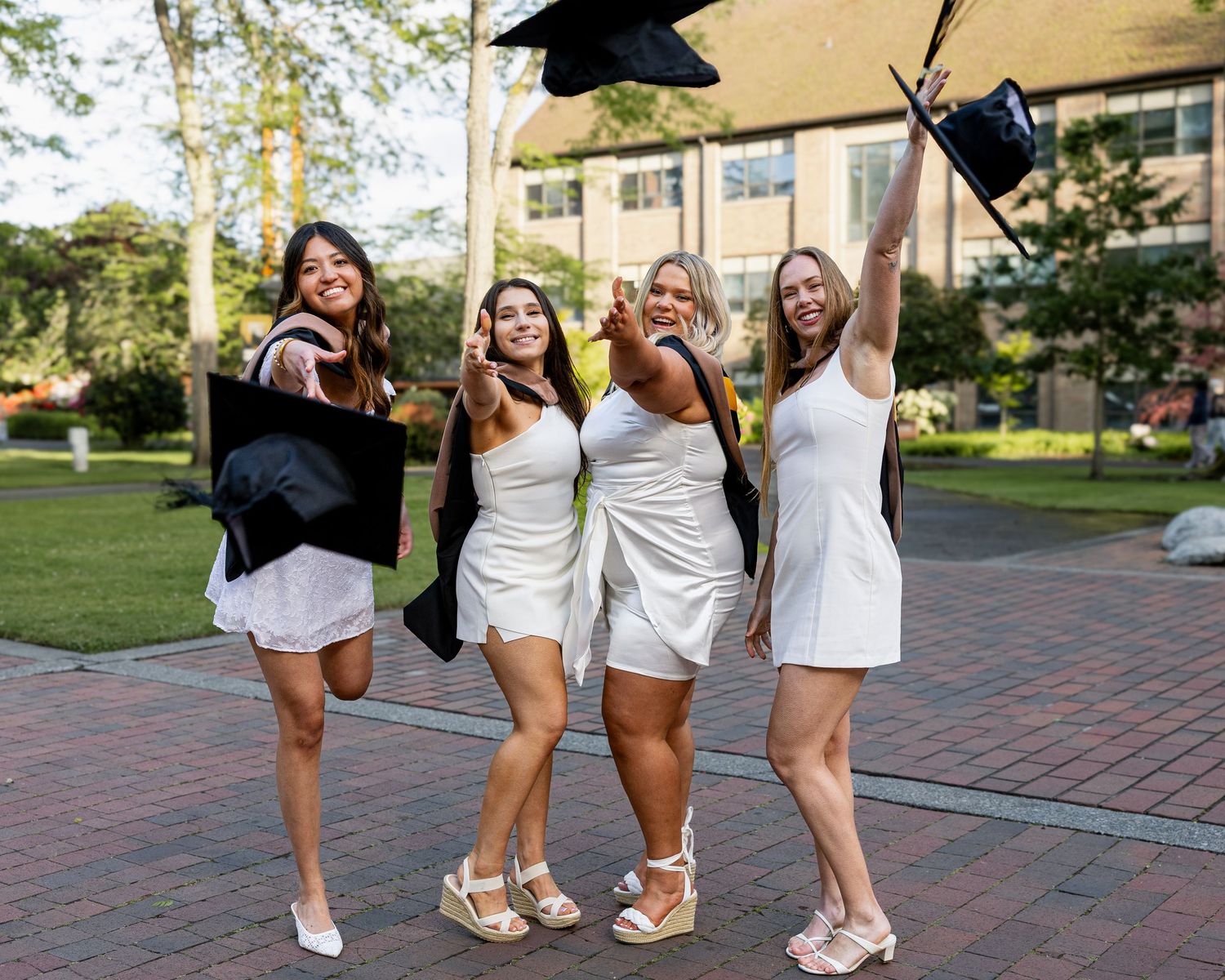 Four young women in white dresses celebrating graduation while tossing caps outdoors on campus.