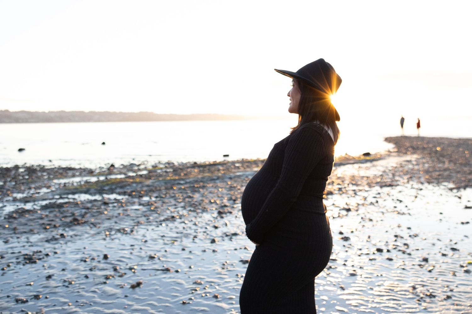 Pregnant woman in black outfit and hat standing on a rocky beach at sunset, enjoying a peaceful moment.