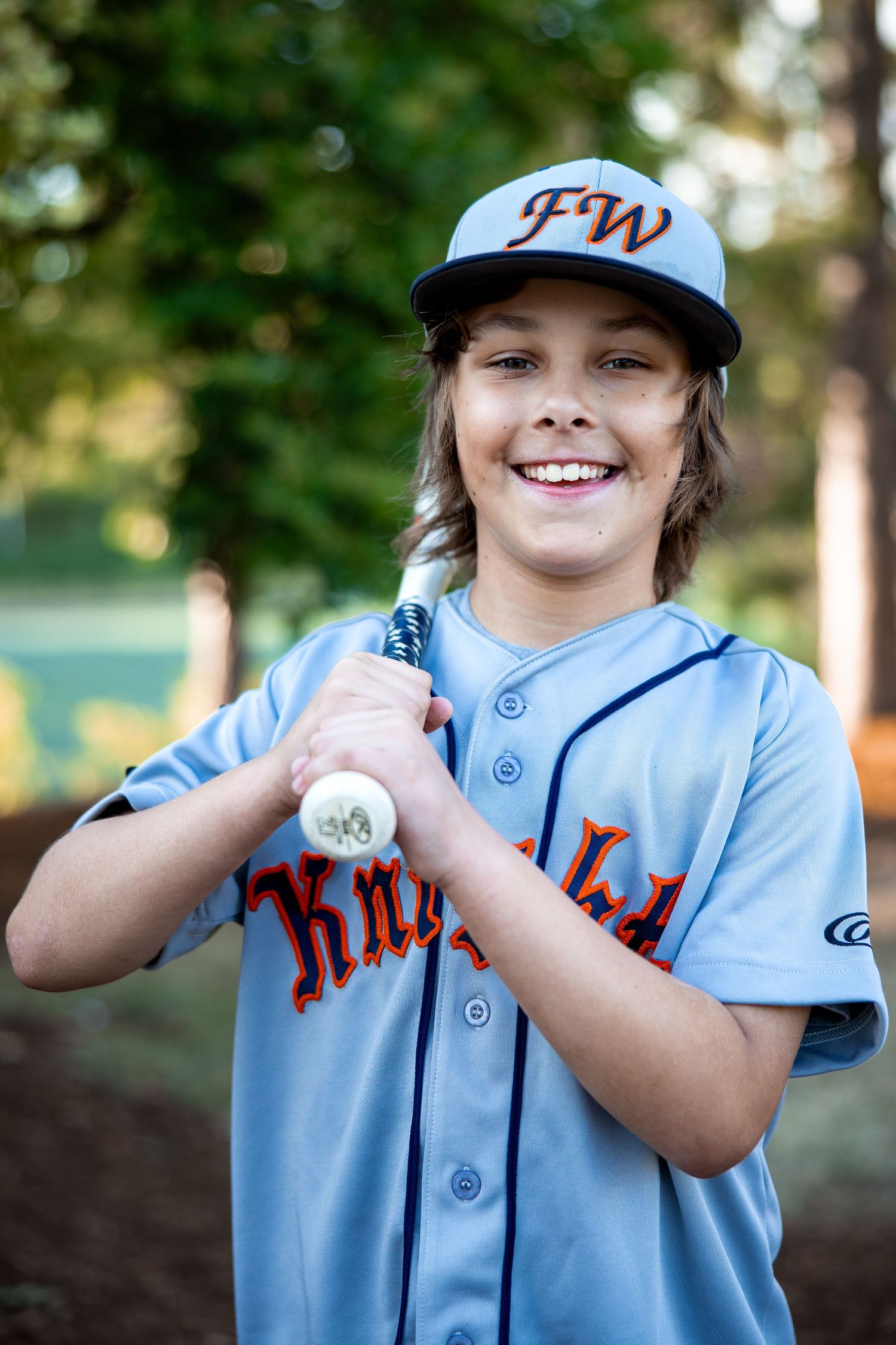 Smiling young player in gray FW Knights baseball uniform holds a bat for an outdoor portrait.