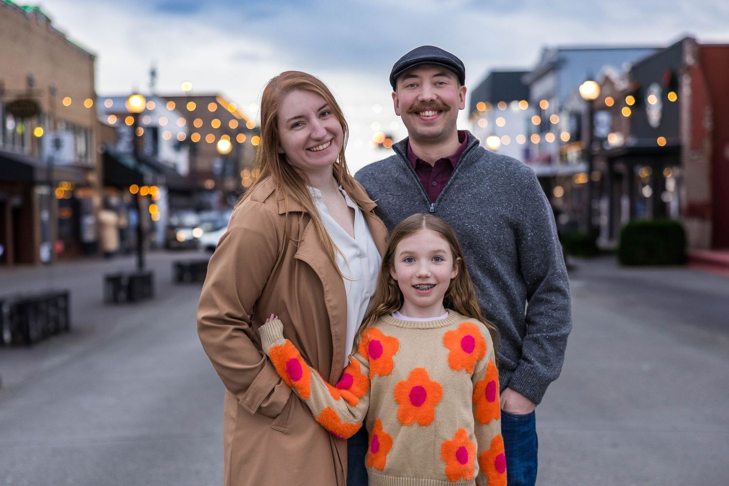 Happy family of three posing on festive lit street at dusk in casual outfits.