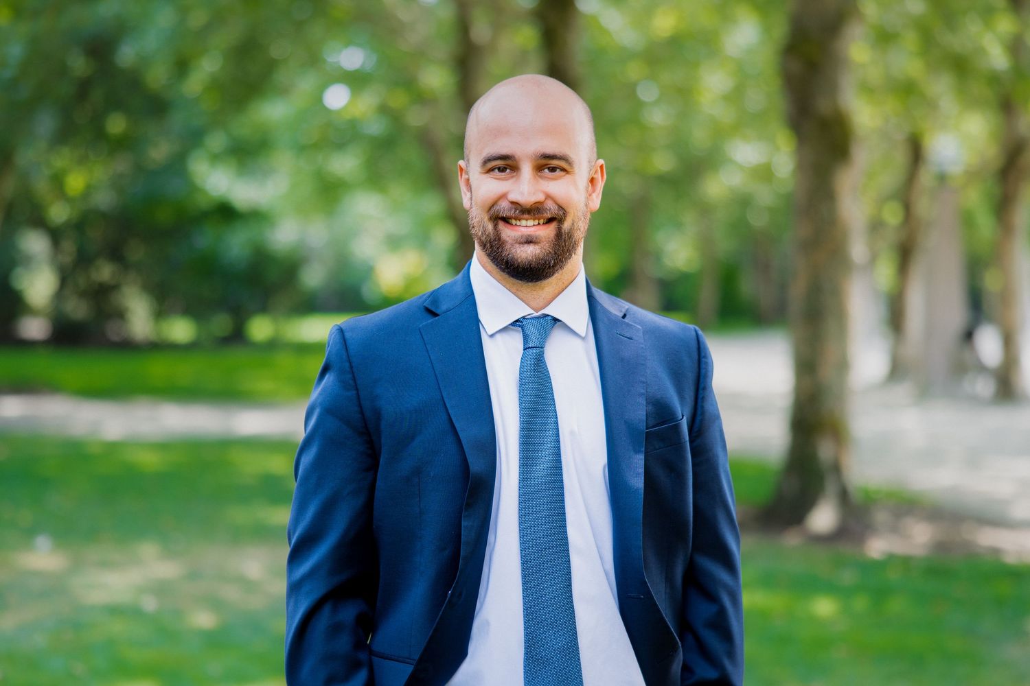 Bald man with beard in navy suit and light blue tie smiling in a green park.