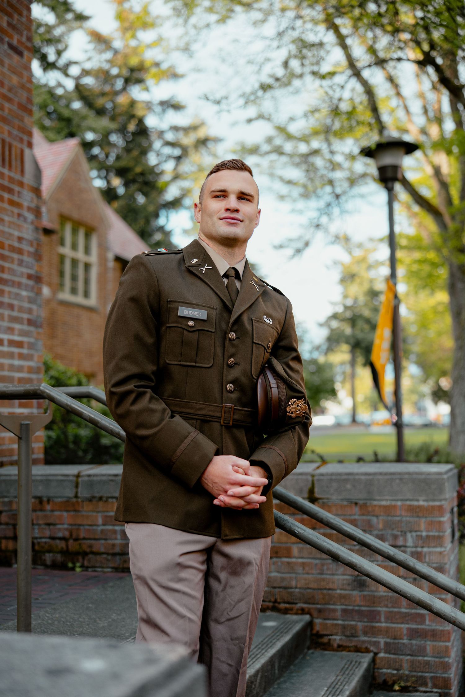 Young man in military dress uniform standing on steps outside a brick building surrounded by trees.