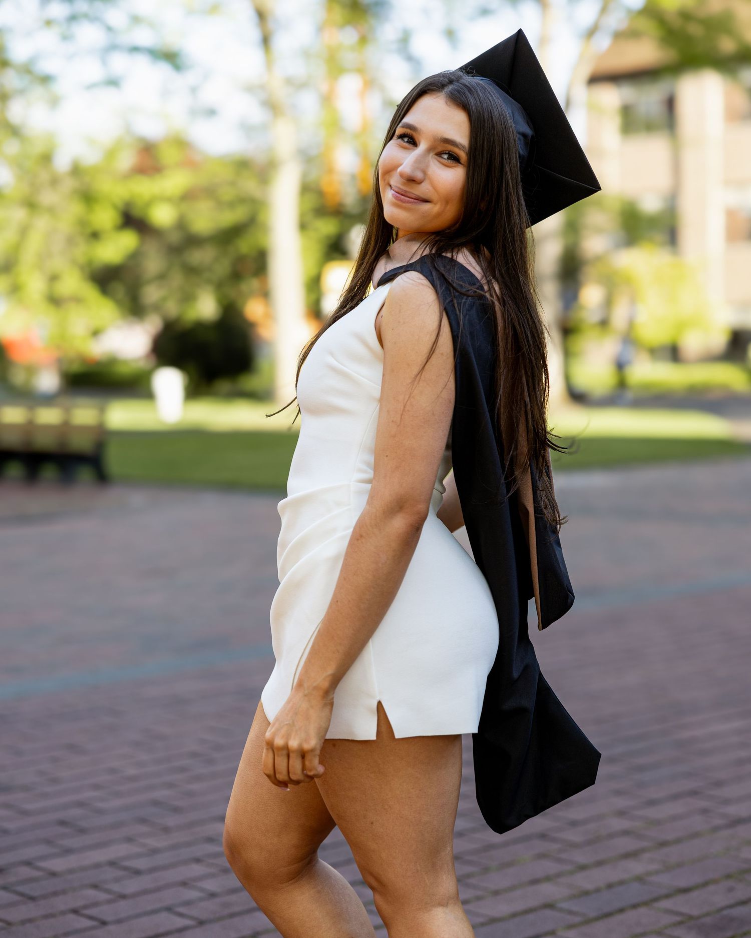 Smiling graduate in white dress holding cap and gown over shoulder on campus.