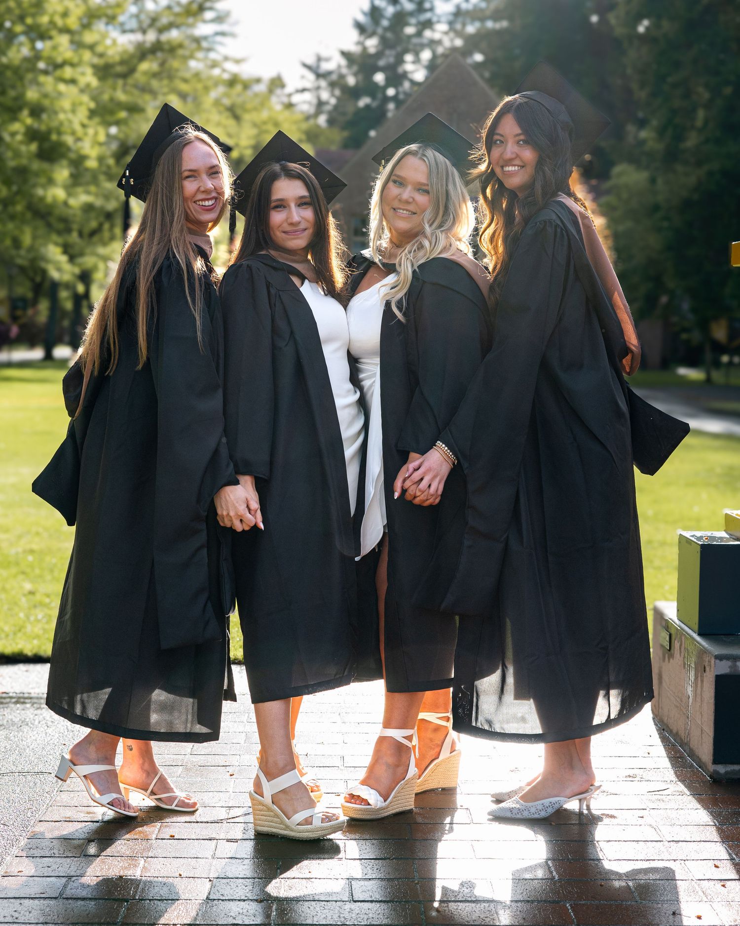 Four female graduates in black caps and gowns posing together outdoors in sunny campus setting.
