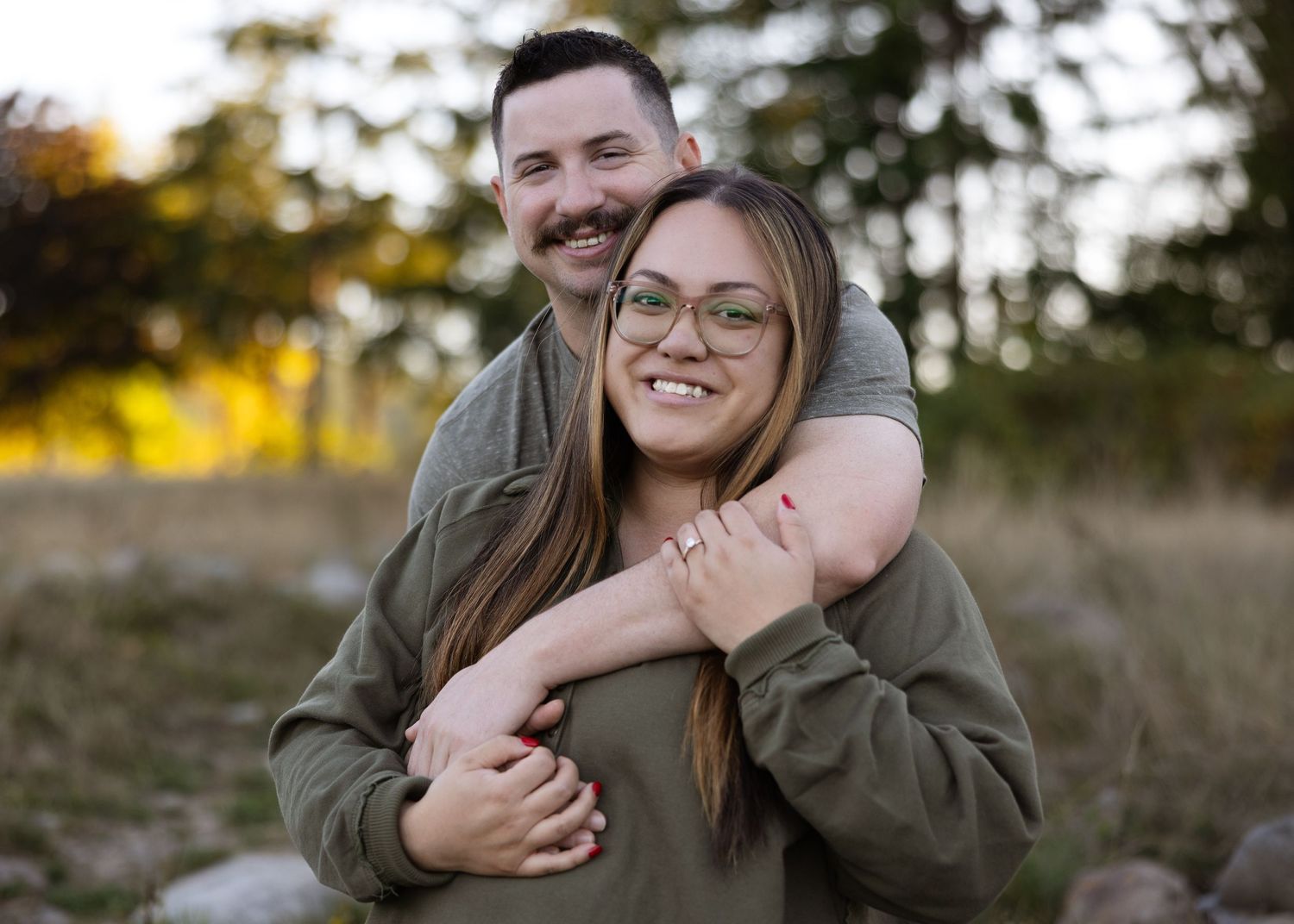 Smiling couple sharing a warm hug outdoors with autumn trees in background.