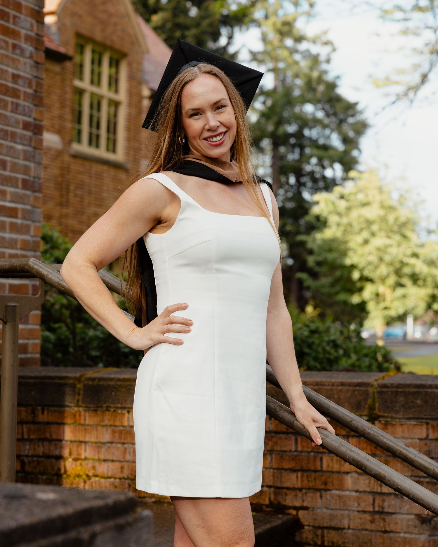 Smiling graduate in white dress and cap posing on brick steps outdoors.