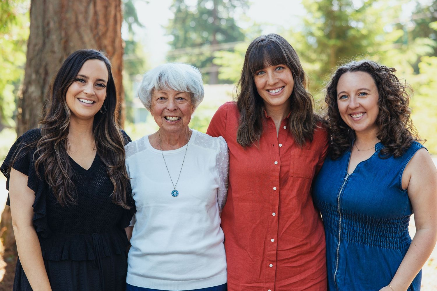 Four women of different generations smiling together outdoors in a sunny park.