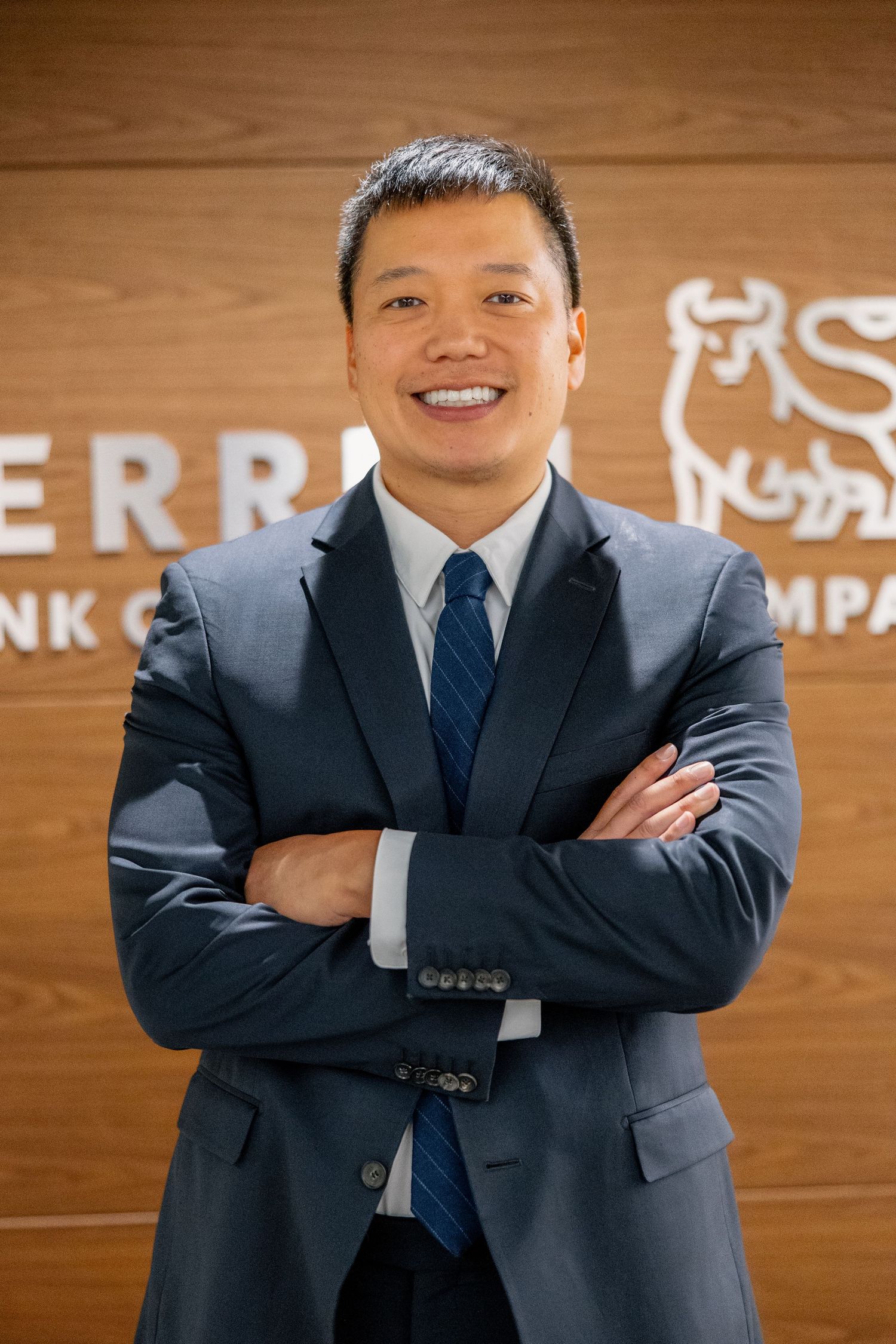 Smiling Asian businessman in navy suit with arms crossed in front of Merrill Lynch sign.