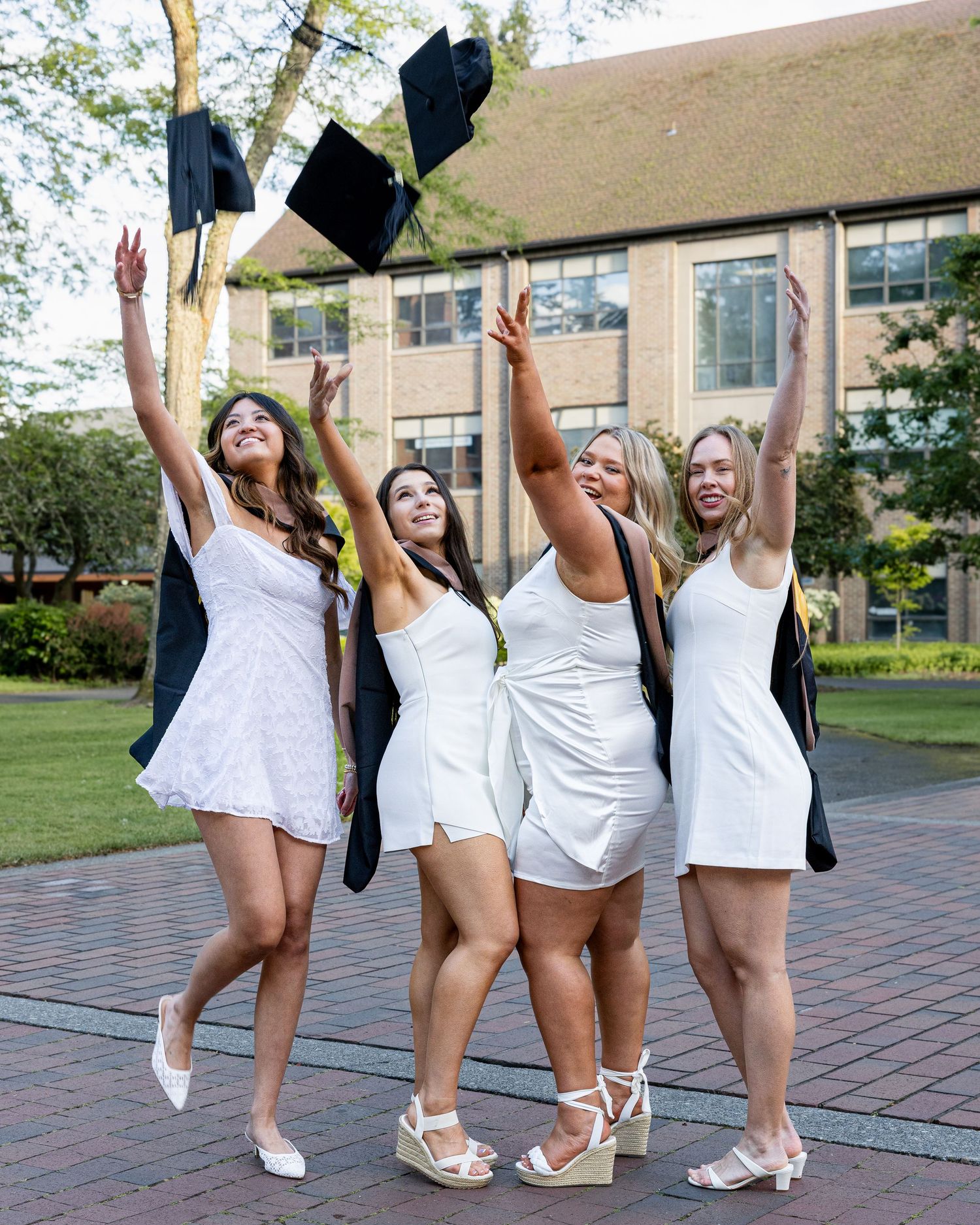 Four young women in white dresses toss graduation caps in the air celebrating their college graduation outdoors.