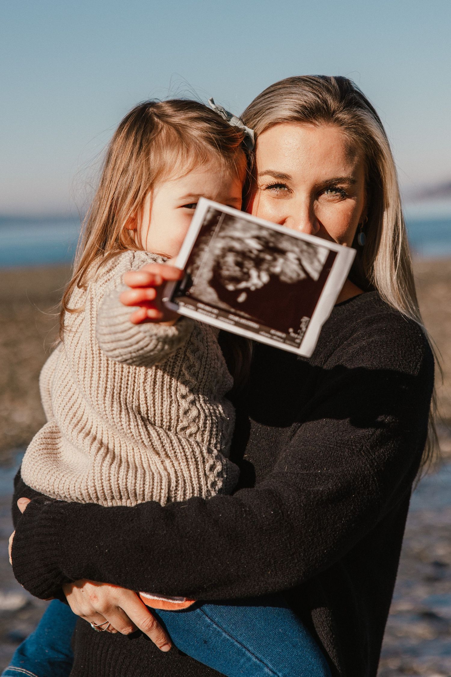 Woman holding toddler girl who displays ultrasound photo on sunny beach, pregnancy announcement.