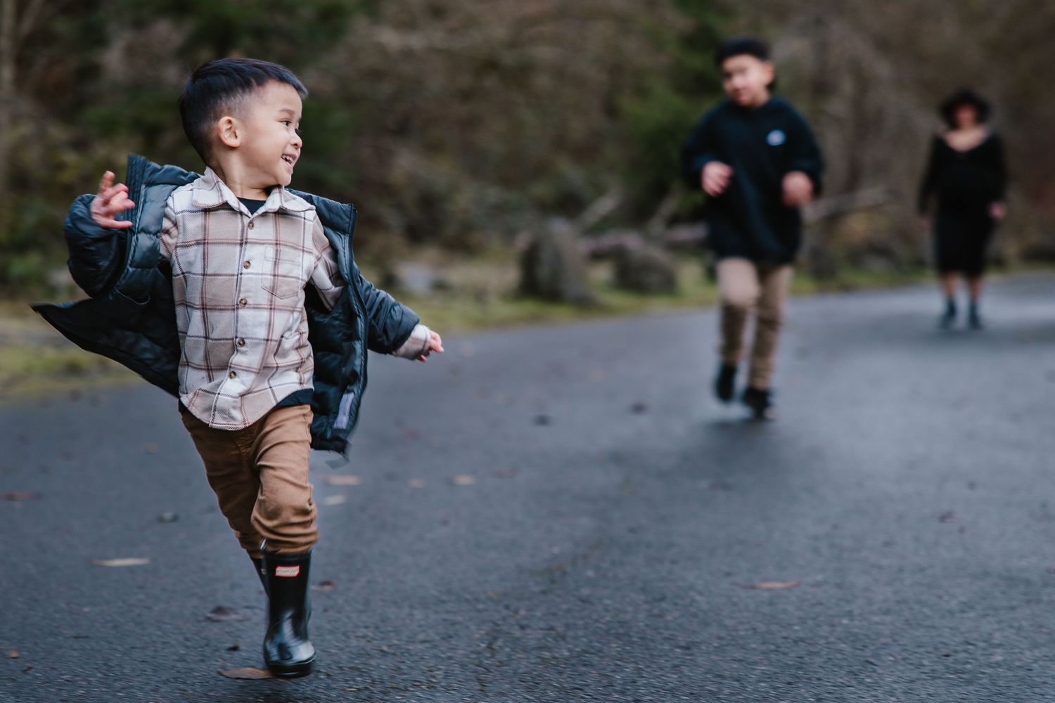 A joyful young boy runs down a path in rain boots while two children walk behind him outdoors.