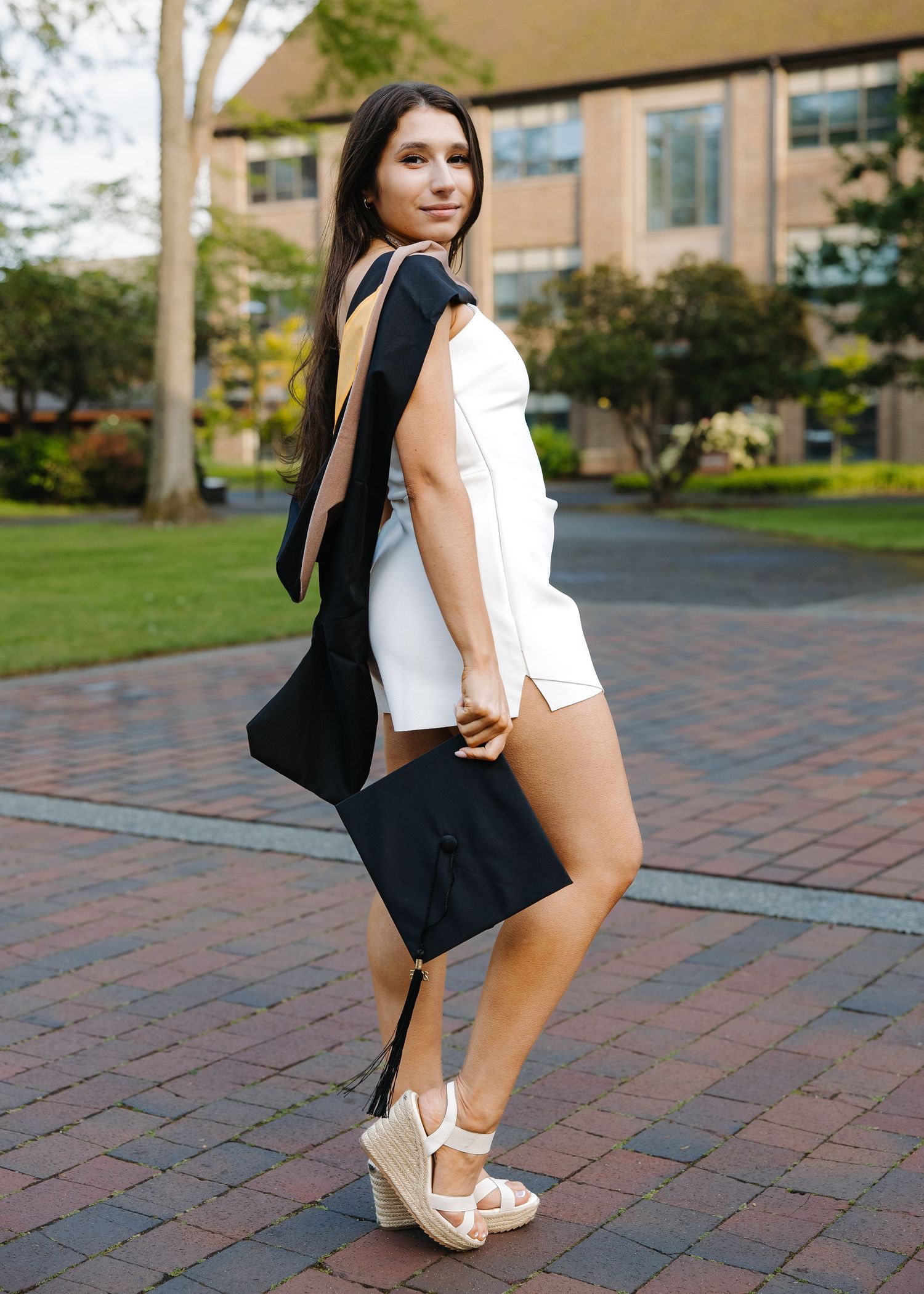 Graduate in white dress carrying cap and gown on sunny brick campus path.