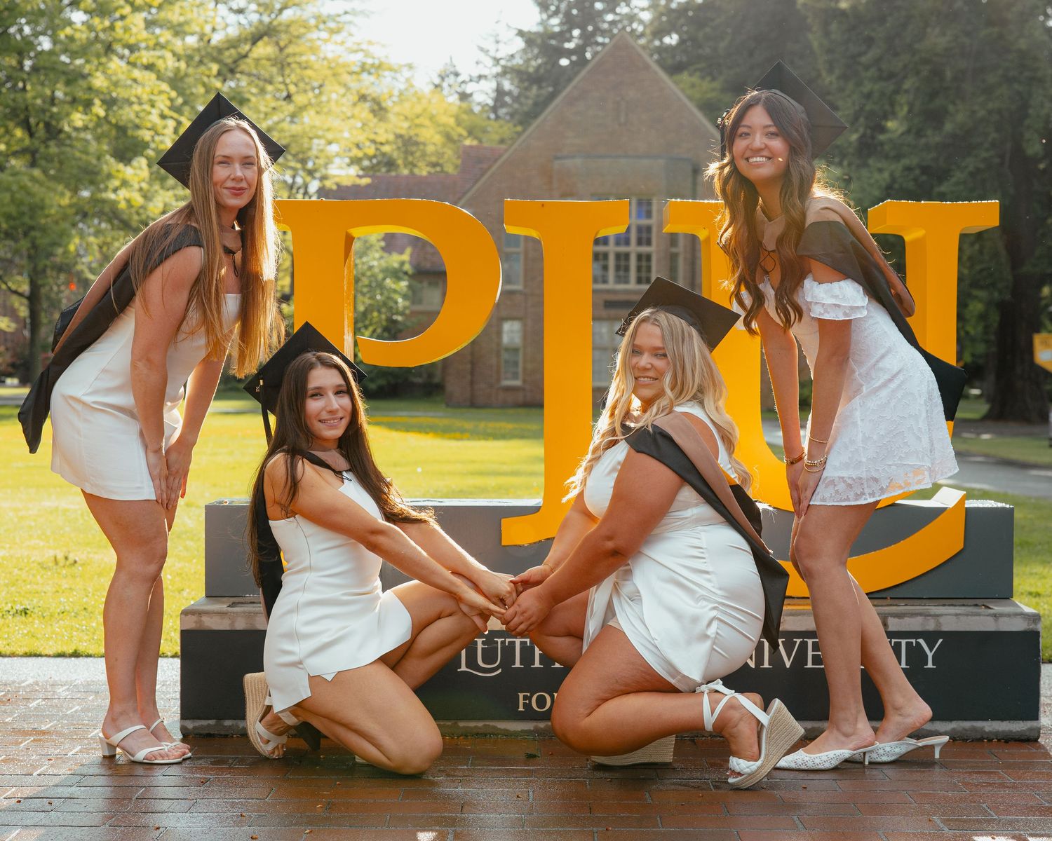 Four female graduates in white dresses posing playfully in front of PLU university sign.