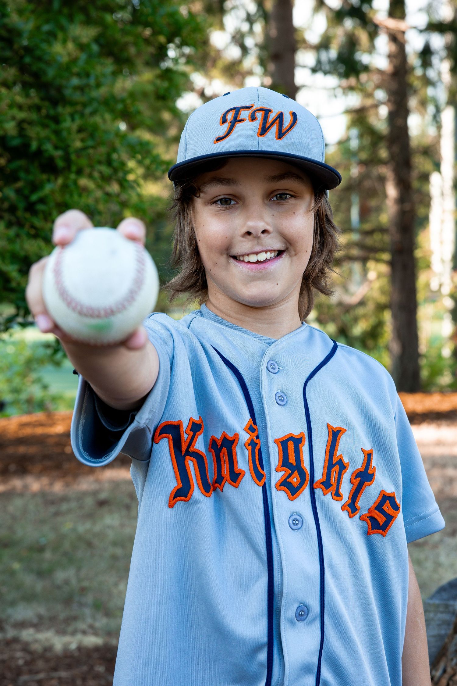 Smiling boy in FW Knights baseball jersey and cap holding a baseball outdoors.