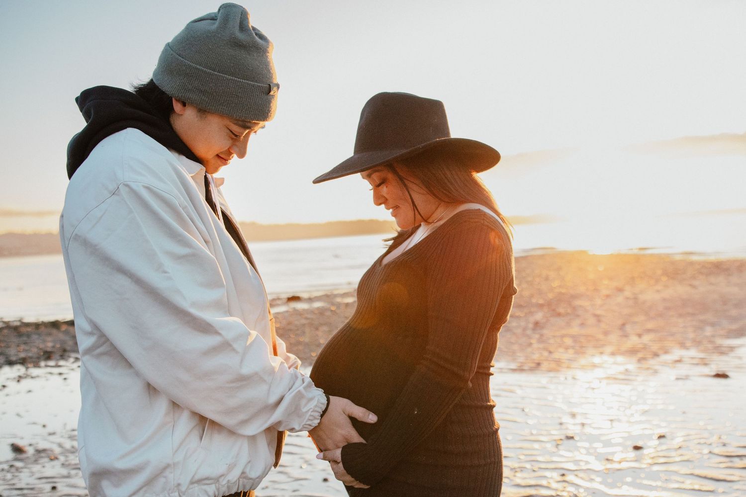 Couple tenderly touching pregnant belly together on a beach during a beautiful sunset.