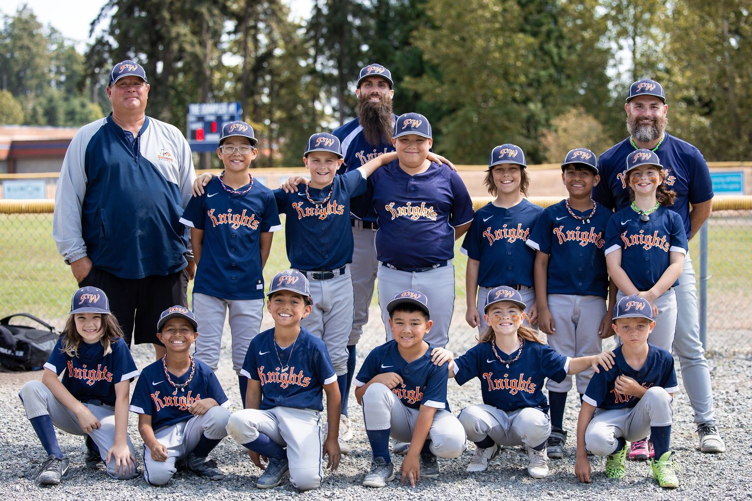 Youth Knights baseball team in navy uniforms poses for a group photo with coaches on a sunny day.