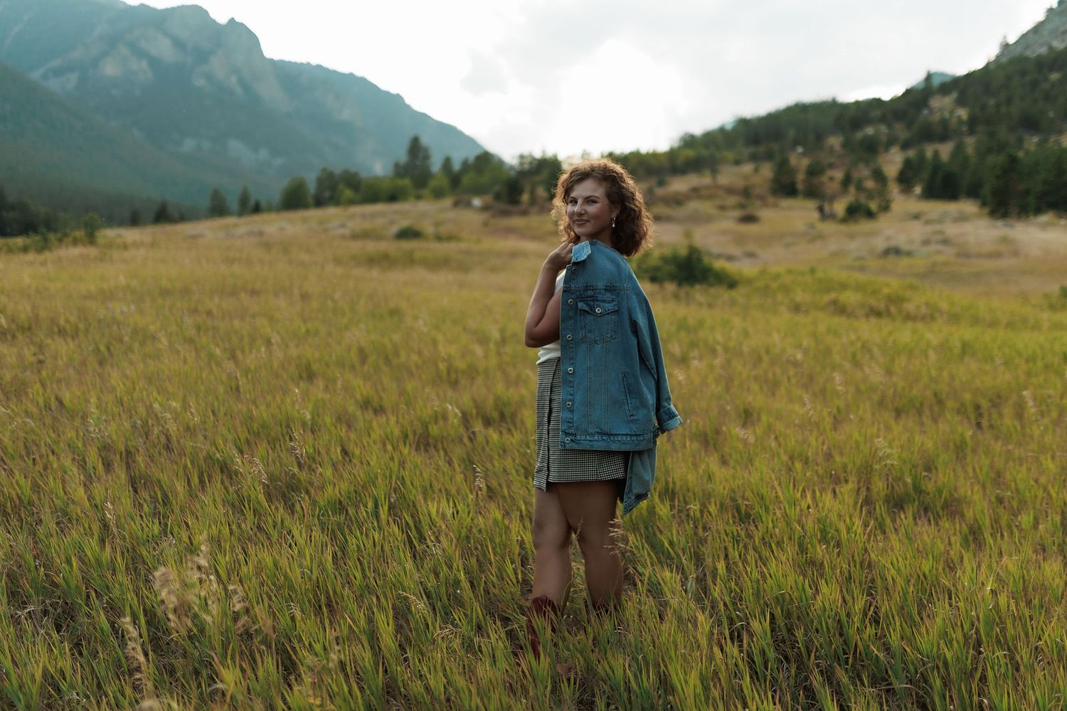 Woman in a denim jacket standing alone in a vast green mountain meadow on a cloudy day.