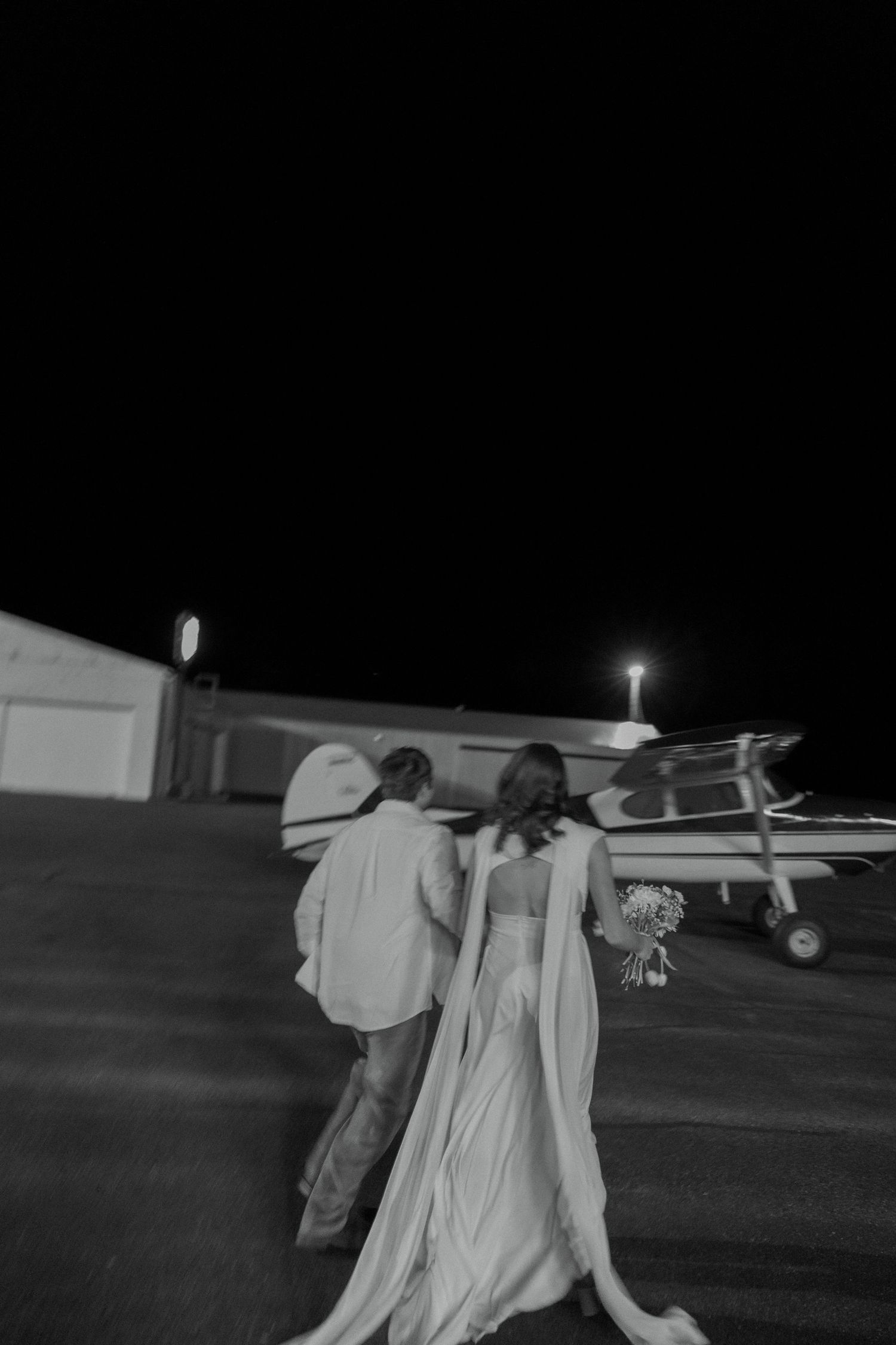 Black and white photo of a bride and groom walking toward a small plane at night, bride holding a bouquet.
