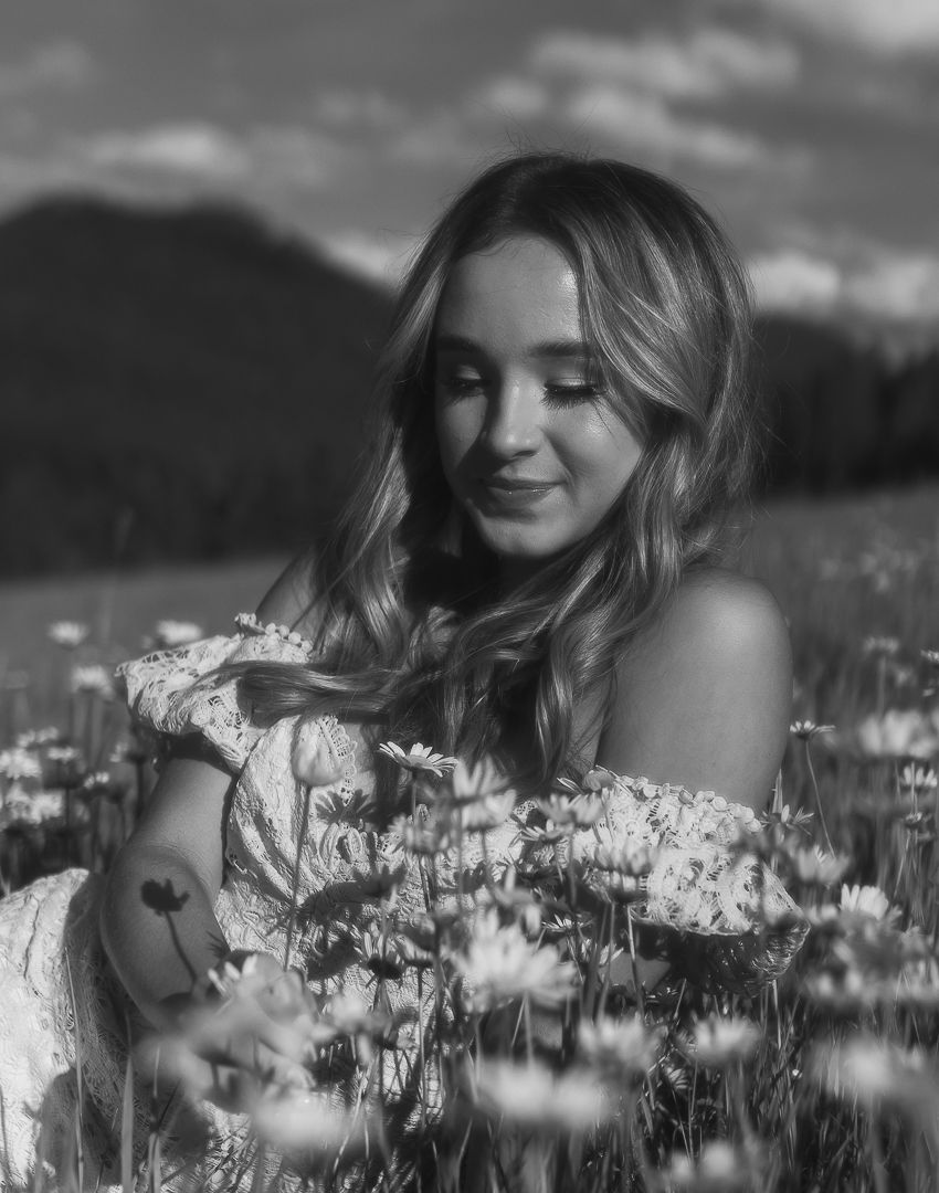 Black and white portrait of a young woman with long hair sitting in a blooming daisy field.