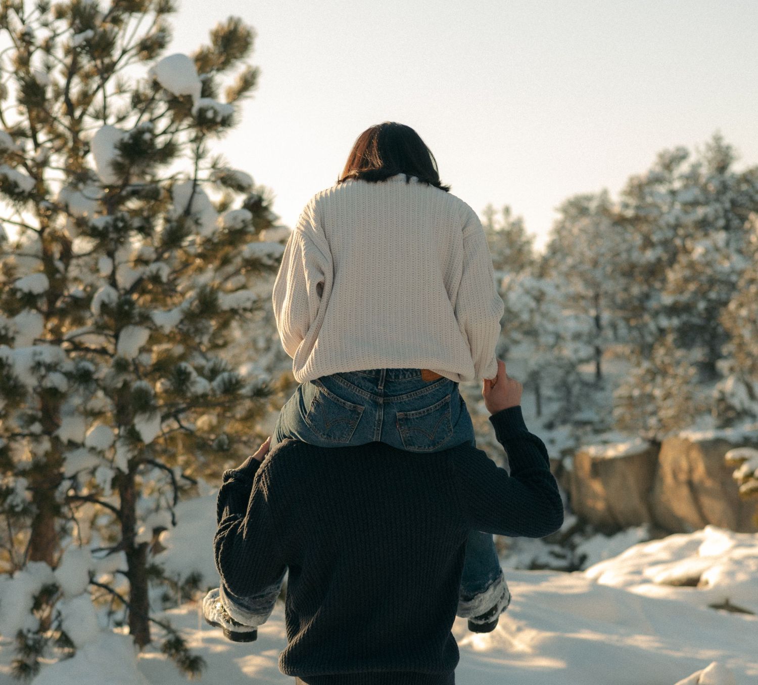 A child sits on an adult's shoulders in a snowy winter forest with snow-covered pine trees.