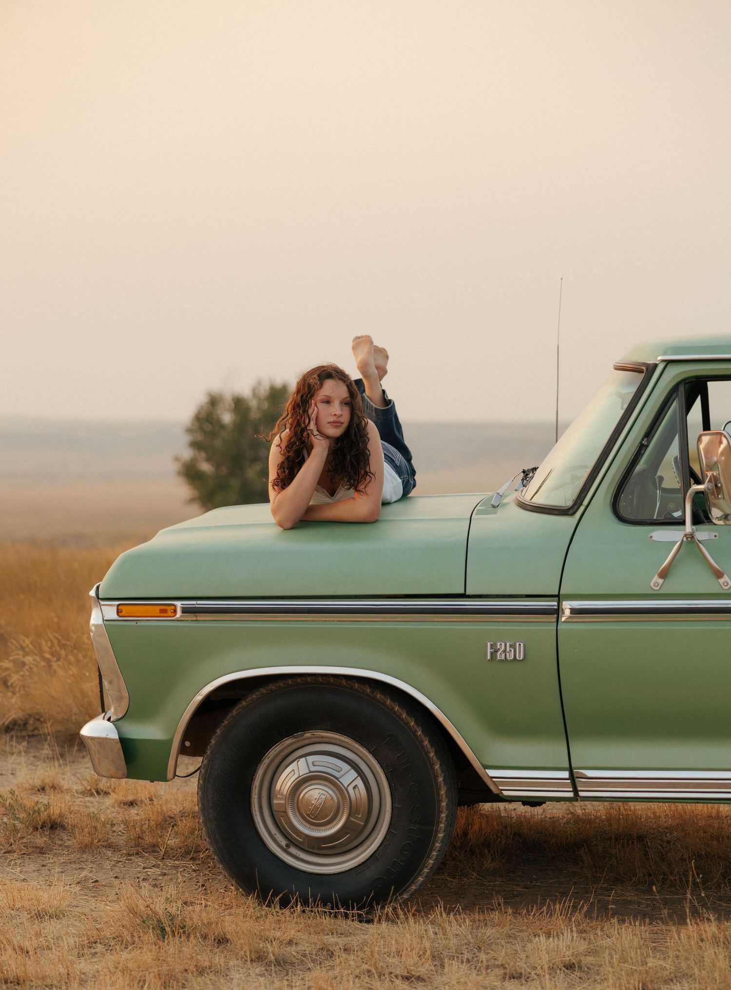 Young woman lying on the hood of a vintage green Ford F-200 truck in a golden field at dusk.