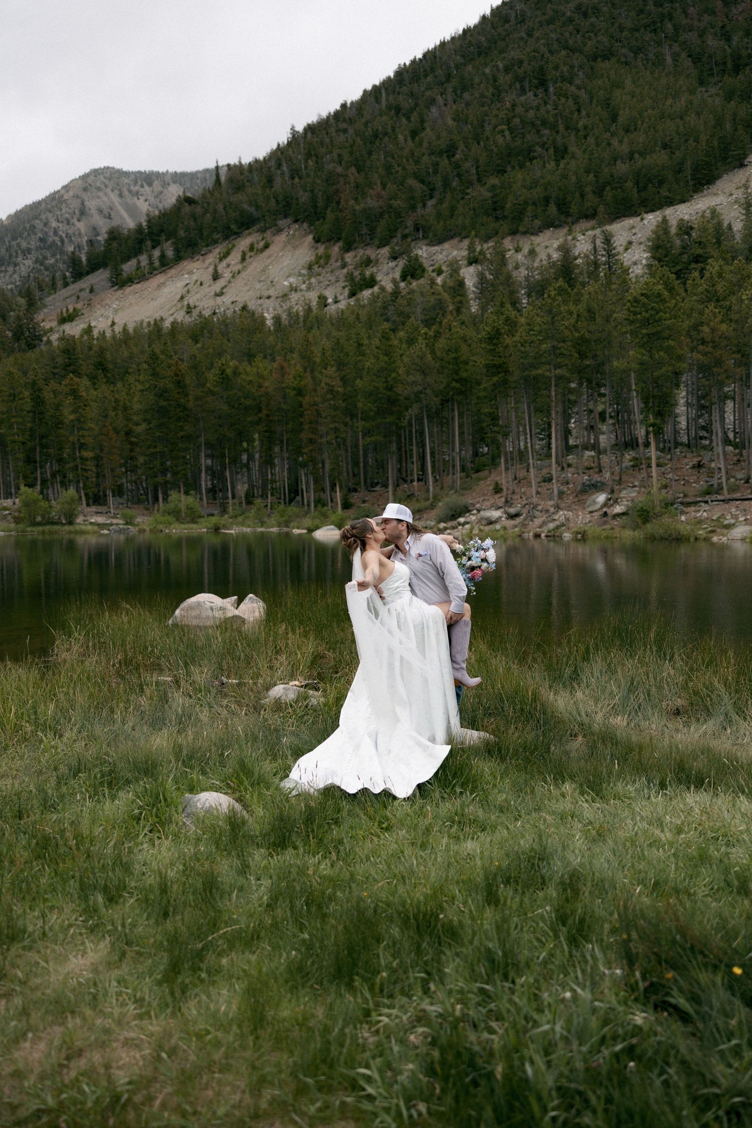Couple sharing a kiss in wedding attire by a mountain lake surrounded by pine trees and lush green grass.