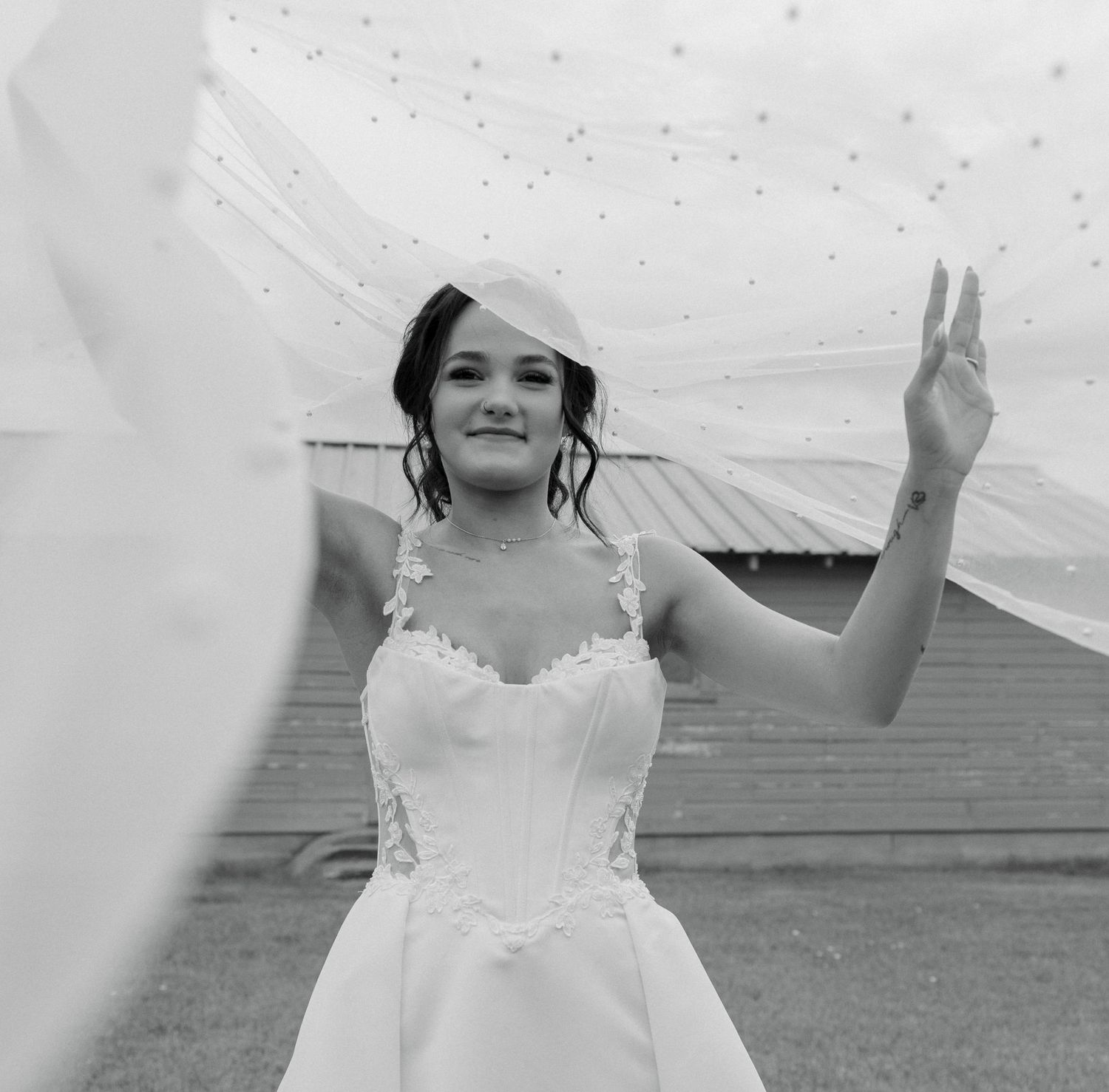 Black and white photo of a smiling bride in a lace wedding dress and veil, posing outdoors with veil flowing in the wind.