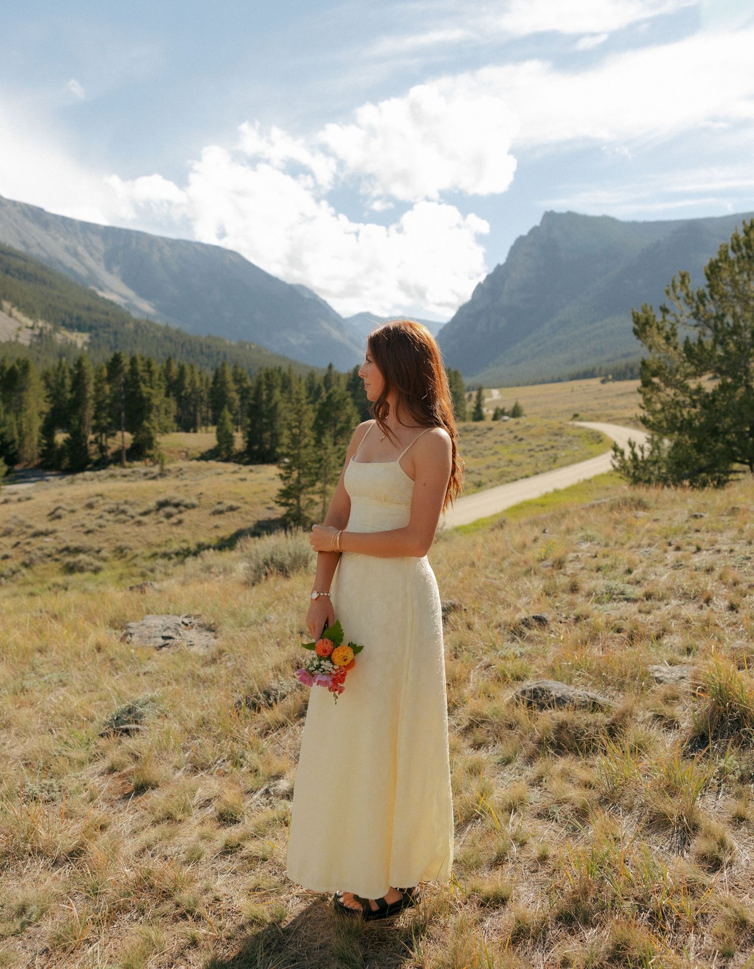 Woman in yellow dress holding wildflowers stands in scenic mountain meadow with pine trees and cloudy sky.